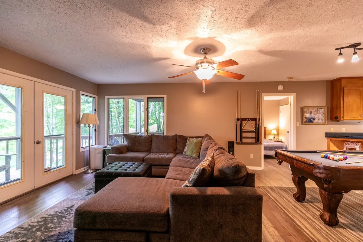 A spacious downstairs recreation room featuring a large sectional sofa positioned near a pool table, with expansive windows that let in natural light. The relaxed setup is ideal for gathering with friends and family, enjoying games, or unwinding in front of the HDTV.