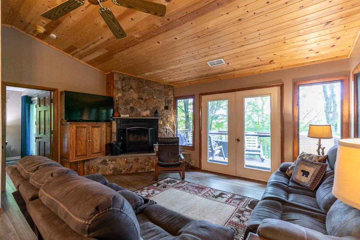 A welcoming living area featuring a stone fireplace, large windows revealing lush greenery outside, and comfortable sofas arranged for conversation or relaxation. A rug anchors the seating area, suggesting a space perfect for gatherings or cozy evenings.