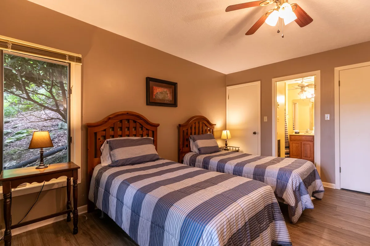 A twin suite is depicted with two twin beds in striped bedding, a nightstand with a lamp between them, and a window framing tranquil greenery outside. Subtle artwork and soft lighting contribute to a quiet, comfortable ambiance perfect for children or friends.