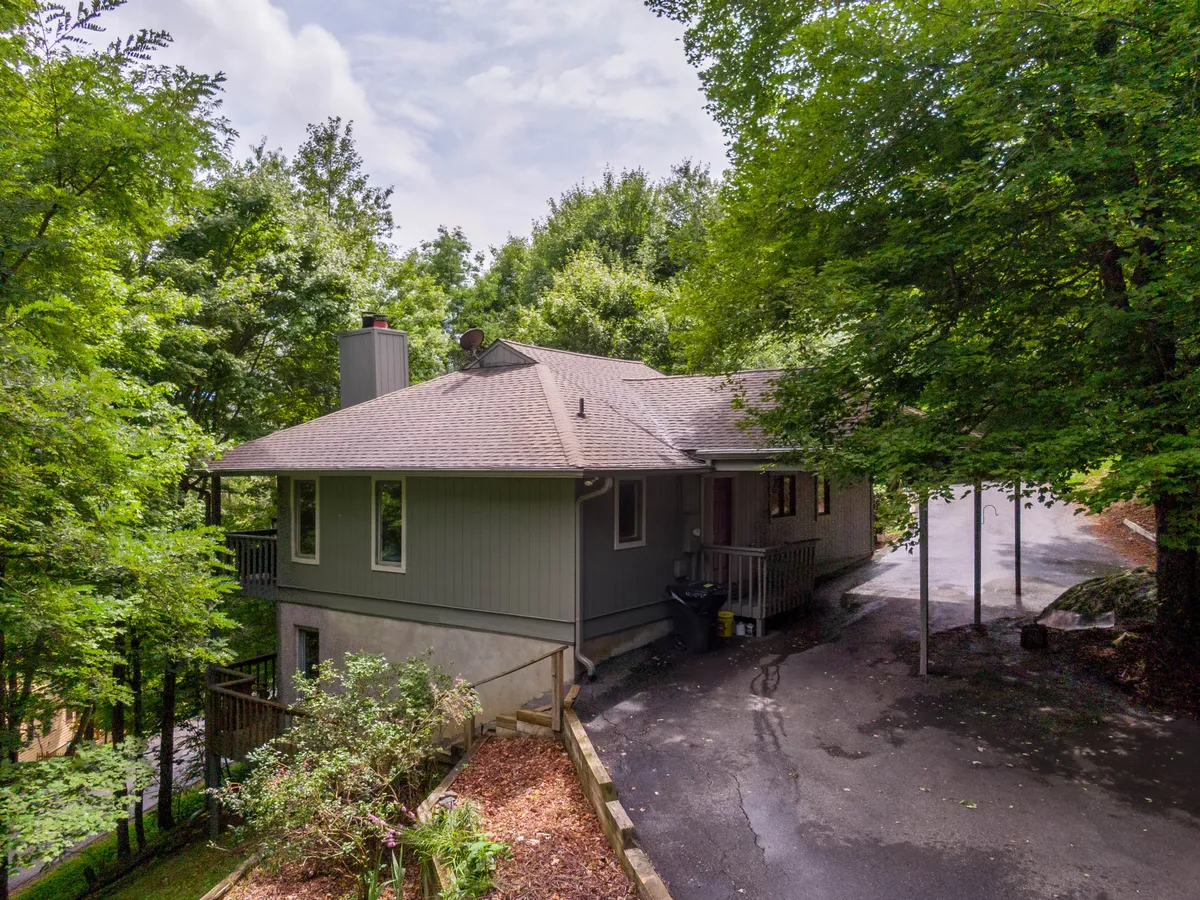 An exterior shot of the house nestled among tall trees, with a welcoming driveway and porch visible. The setting exudes tranquility and direct access to nature, emphasizing the home's connection to its wooded surroundings.