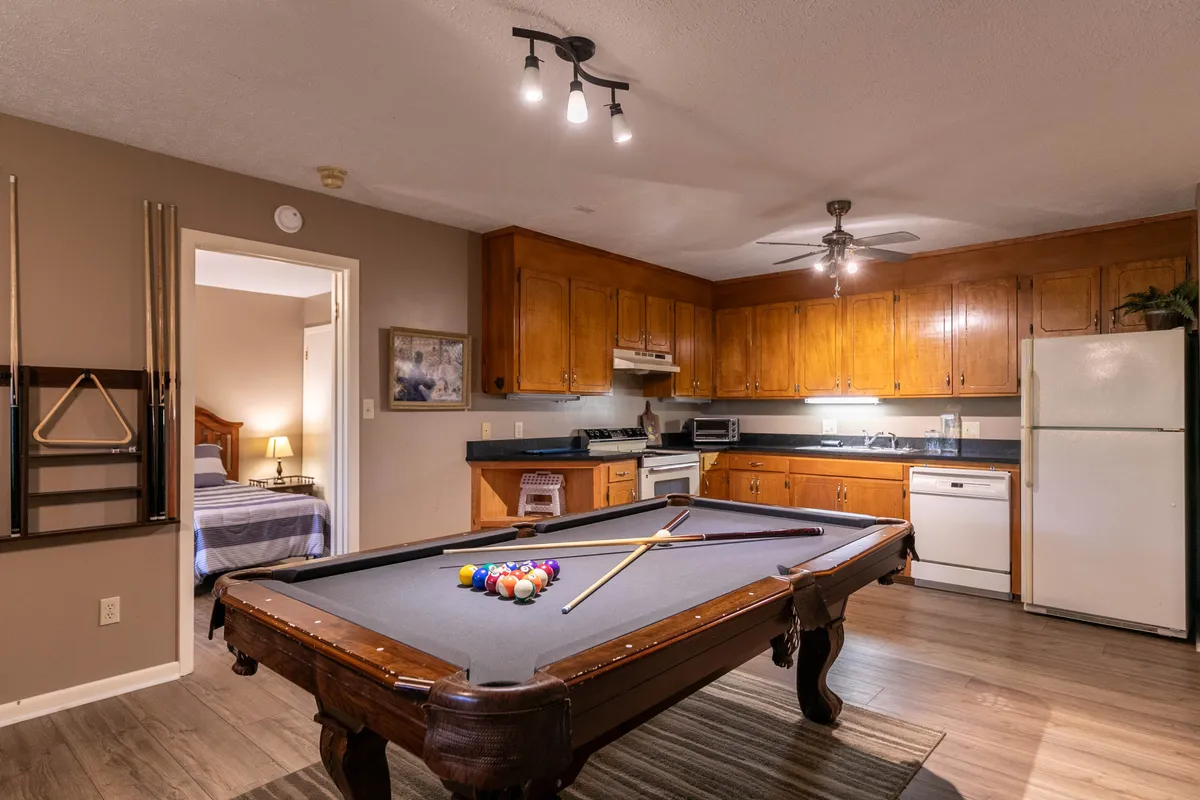 A view of the downstairs game area, highlighting the pool table in the foreground and a kitchenette with cabinetry and a refrigerator in the background. The space is arranged for both recreation and convenience, with easy access to snacks or drinks.