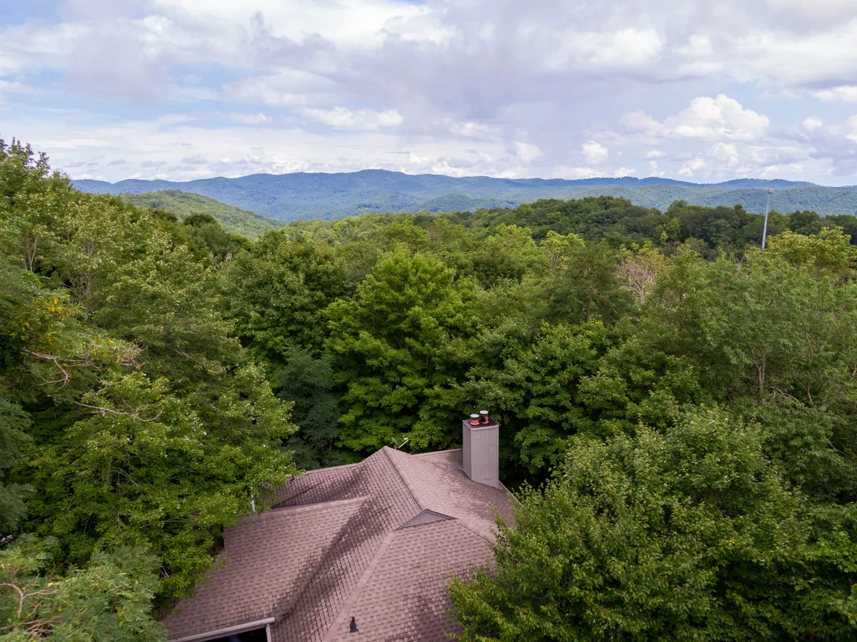 This image features the roof of the home nestled amid tall trees with distant mountain peaks visible in the background. The overall palette is cool, evoking a crisp and serene mountain atmosphere. The elevated vantage point hints at panoramic winter views, aligning perfectly with the section's emphasis on the home’s outlook and proximity to mountain adventures.