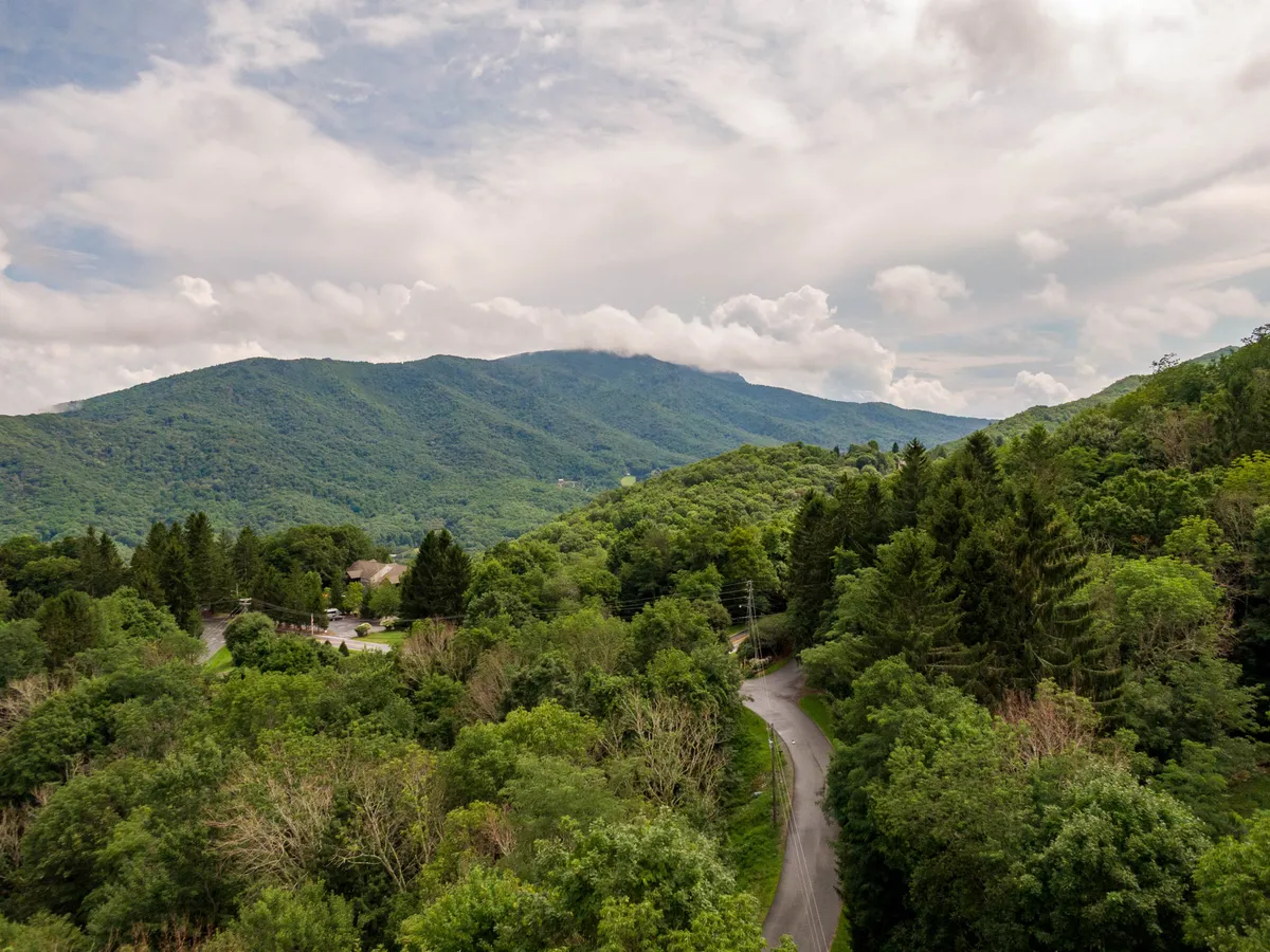 A sweeping view of majestic mountains with a winding road below, surrounded by dense greenery. The scene suggests endless opportunities for hiking, scenic drives, and exploring the natural beauty of the region. The lighting hints at either early morning or late afternoon, ideal times for outdoor adventure.