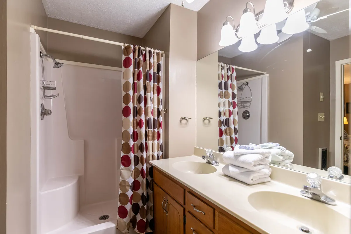 A bright, functional en-suite bathroom with a double sink, large mirror, and a decorative shower curtain over the shower area. Ample space and thoughtful details create an inviting setting for morning routines and relaxation.