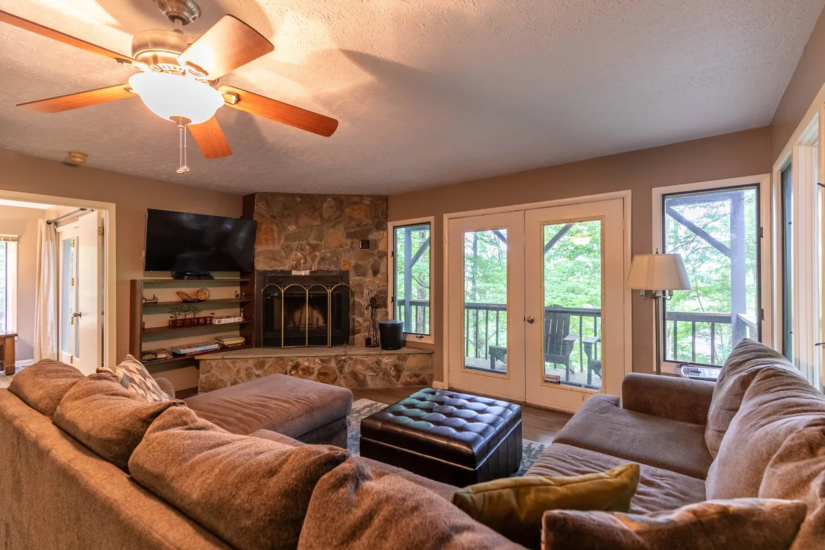 This image shows a spacious living area featuring a large sectional sofa facing a striking stone fireplace. The room is bathed in natural light from expansive windows, which offer scenic views. The cozy atmosphere is enhanced by a ceiling fan and warm decor, with the living space arranged for relaxation and social gatherings.