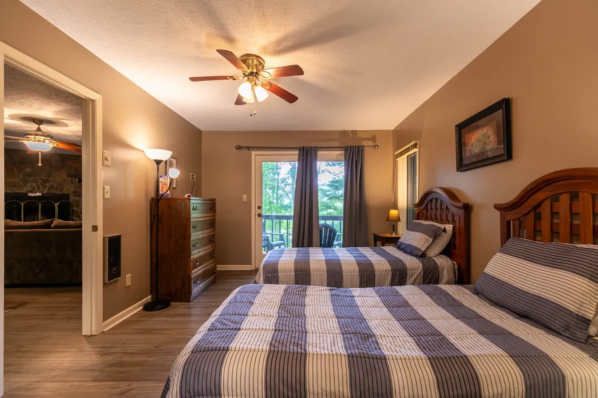 A comfortable bedroom setup with two beds, a dresser, and a window dressed with curtains. The overall atmosphere is homely, and the view outside hints at greenery, suggesting proximity to nature and possibly a shared balcony or porch.