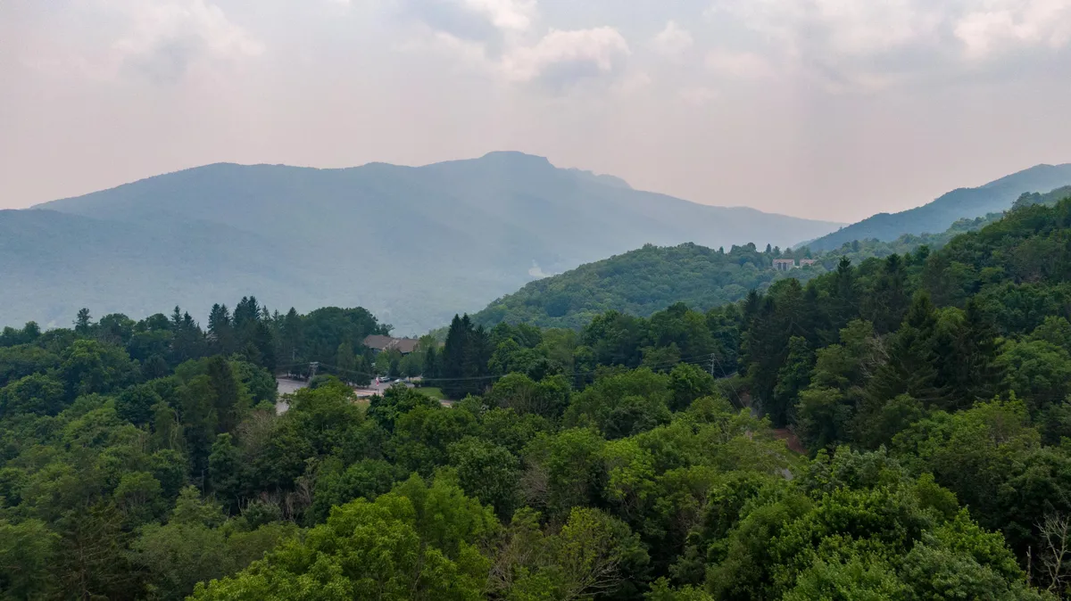 A stunning vista of distant mountains framed by dense greenery and a soft, hazy sky. The scene captures the grandeur of Grandfather Mountain and the surrounding landscape, inviting viewers to imagine the changing beauty across seasons, from snowy mornings to golden autumn.